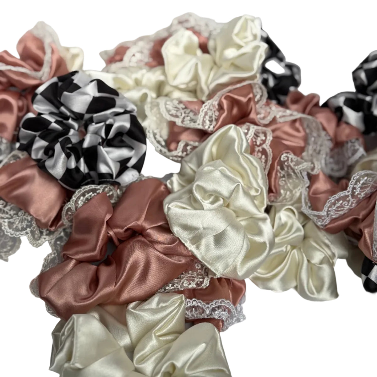 A pile of satin hair scrunchies in black and white checkered, blush pink, and cream satin, some decorated with white lace trim, arranged on a white surface.