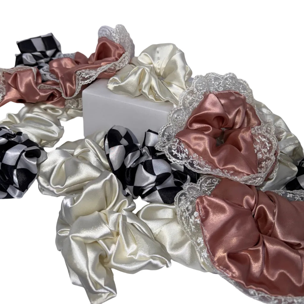 A collection of satin hair scrunchies in various colors and patterns, including black and white checkered, solid cream, and dusty rose with lace trim, arranged on a white surface.