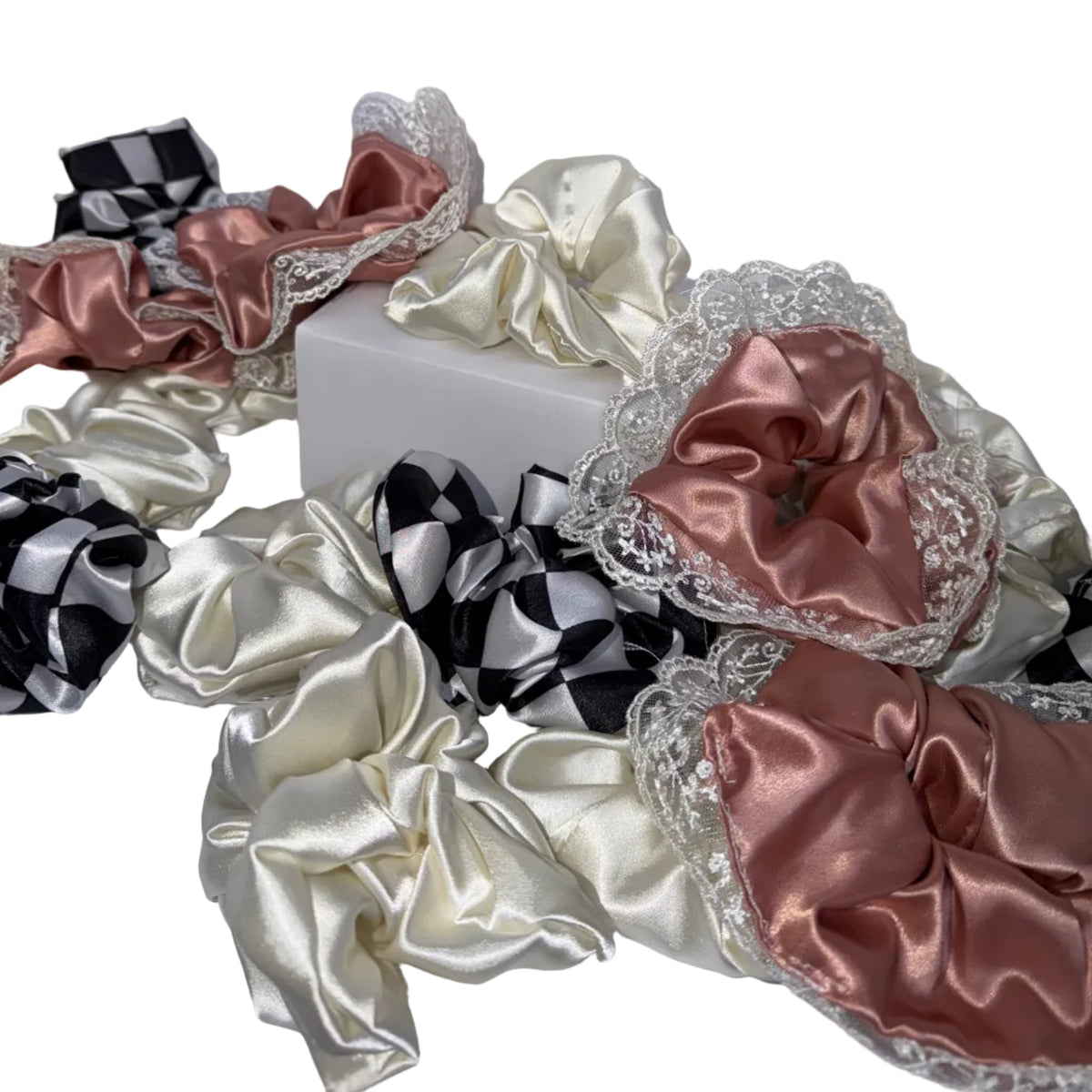 A collection of satin hair scrunchies in various colors and patterns, including black and white checkered, solid cream, and dusty rose with lace trim, arranged on a white surface.